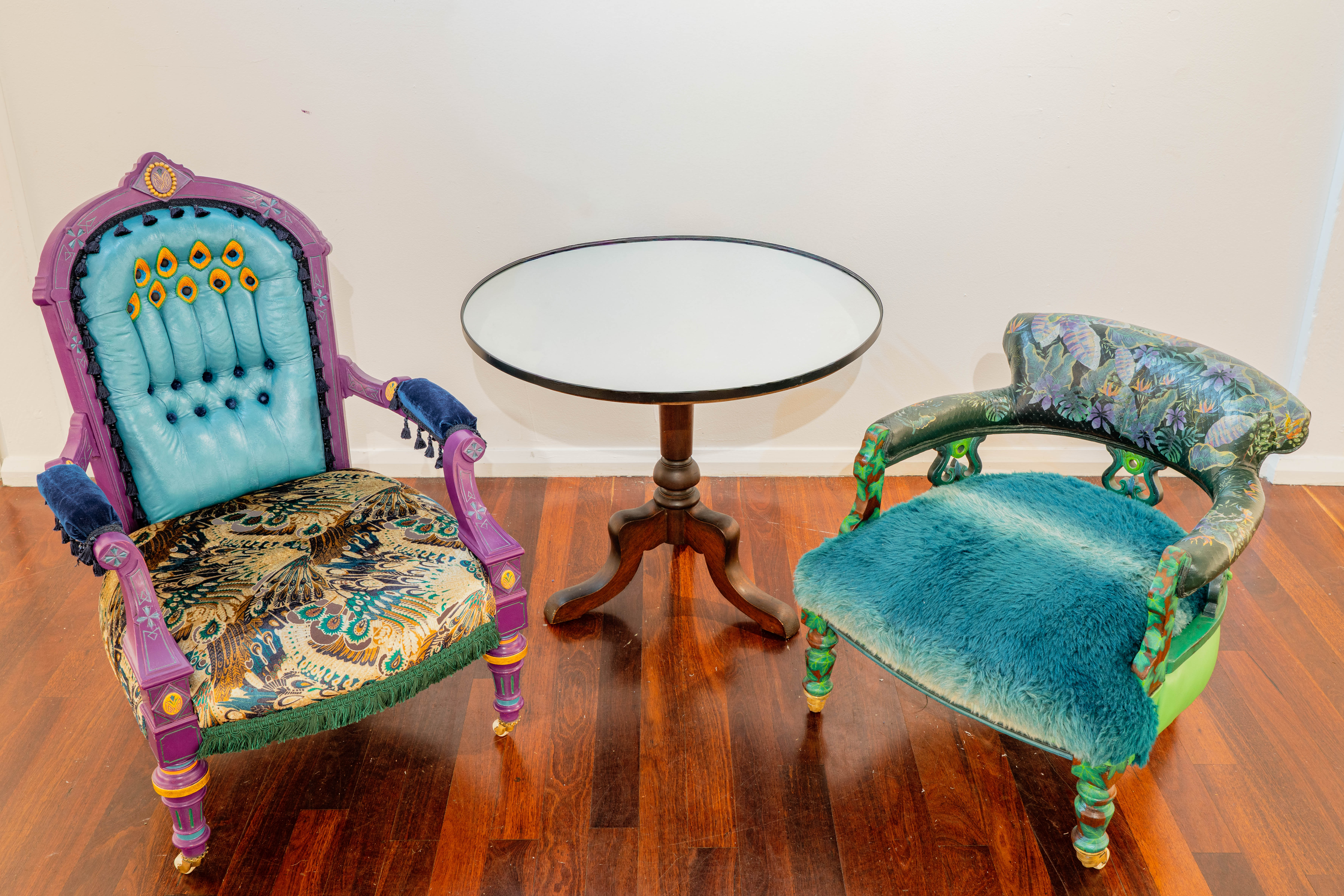 Two antique chairs and a side table, one chair decorated with a peacock themed colours and fabric and the other a jungle-themed chair with multiple shades of green.